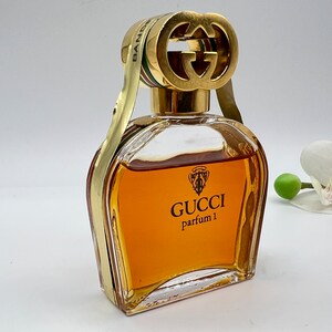 May include: A vintage Gucci perfume bottle with a gold cap featuring the iconic interlocking G logo. The bottle is filled with a golden-amber liquid and has the text "GUCCI parfum 1" on the front.