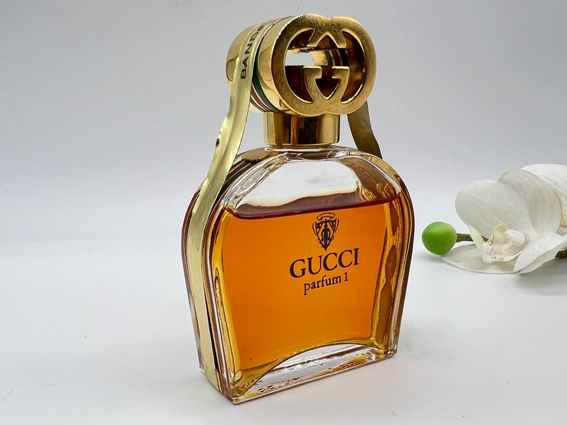 May include: A vintage Gucci perfume bottle with a gold cap featuring the iconic interlocking G logo. The bottle is filled with a golden-amber liquid and has the text "GUCCI parfum 1" on the front.