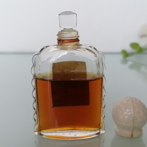 May include: A clear glass bottle with a textured design and a brown liquid inside. The bottle has a clear glass stopper and a brown label. The bottle is sitting on a white surface.