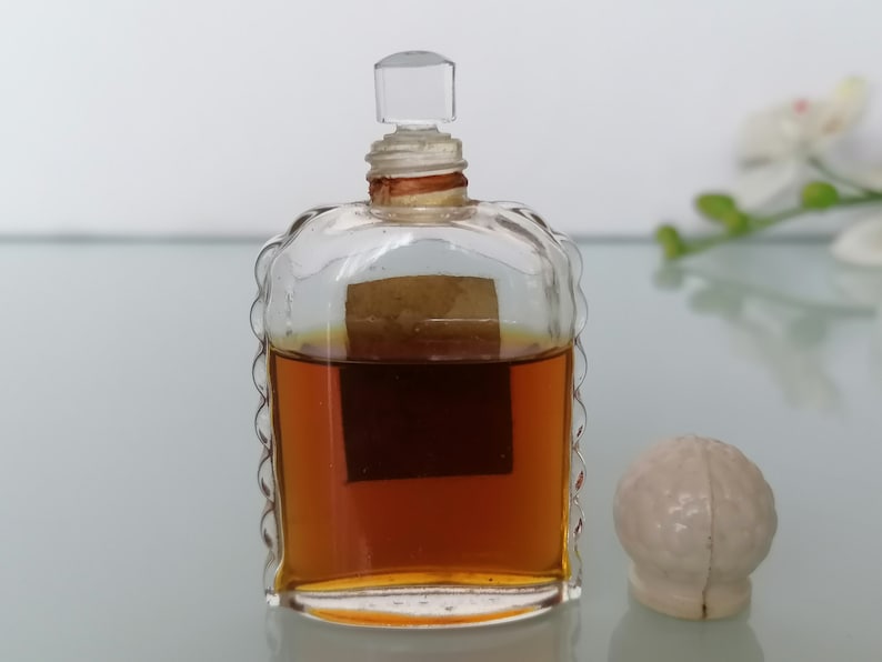 May include: A clear glass bottle with a textured design and a brown liquid inside. The bottle has a clear glass stopper and a brown label. The bottle is sitting on a white surface.