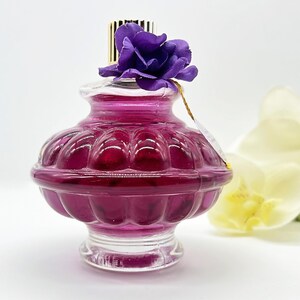 May include: A decorative, clear glass bottle with a deep magenta liquid inside. The bottle has a bulbous shape with a ridged design and is topped with a purple fabric flower. A gold-colored label hangs from the bottle's neck.