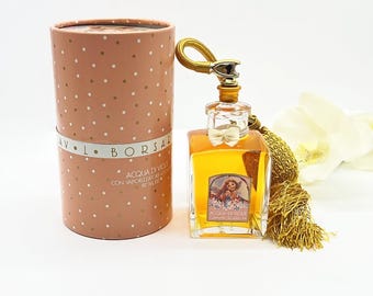 Puede incluir: Un frasco de perfume vintage con una borla dorada y una caja cilíndrica a juego. El frasco de perfume es cuadrado con una tapa dorada y un pequeño lazo. La caja es rosa con lunares blancos y el texto "AV. L. BORSA".