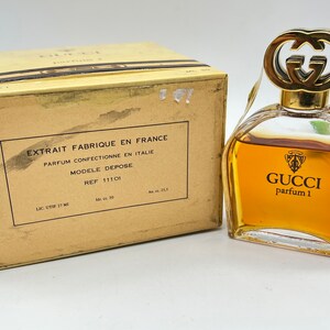 May include: A vintage Gucci perfume bottle with a gold cap and a clear glass body. The bottle is filled with a golden liquid and has the text "GUCCI parfum 1" printed on the front. The bottle is sitting on a white surface.