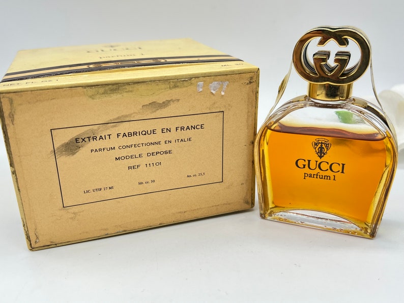 May include: A vintage Gucci perfume bottle with a gold cap and a clear glass body. The bottle is filled with a golden liquid and has the text "GUCCI parfum 1" printed on the front. The bottle is sitting on a white surface.