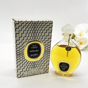 May include: A gold and white box with a black and white geometric pattern. The box contains a clear glass bottle of Mitsouko Eau de Toilette by Guerlain. The bottle is shaped like a teardrop and has a gold cap. The bottle is labeled with the text "Eau de Toilette Mitsouko Guerlain Paris 50 ml".