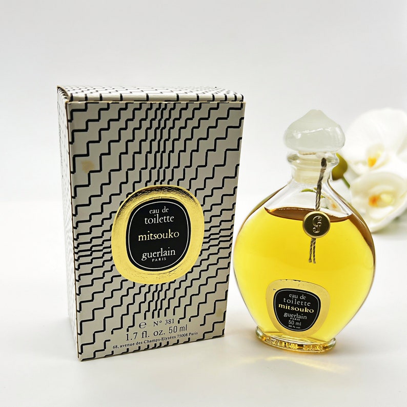 May include: A gold and white box with a black and white geometric pattern. The box contains a clear glass bottle of Mitsouko Eau de Toilette by Guerlain. The bottle is shaped like a teardrop and has a gold cap. The bottle is labeled with the text "Eau de Toilette Mitsouko Guerlain Paris 50 ml".