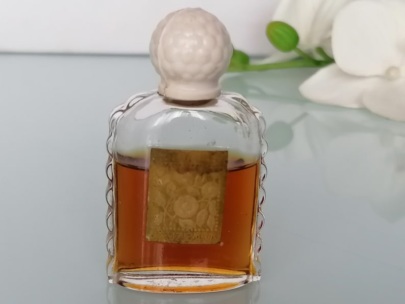 May include: A small, clear glass bottle with a white plastic stopper. The bottle contains a brown liquid and has a gold label with a floral design.