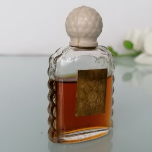 May include: A small, clear glass bottle with a white plastic stopper. The bottle is filled with a brown liquid and has a gold label with a floral design.