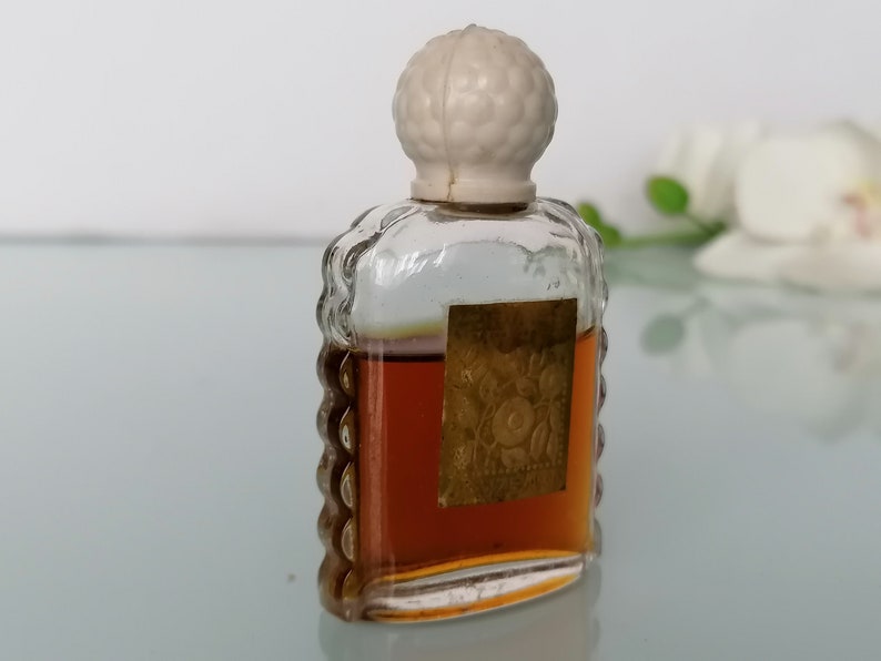 May include: A small, clear glass bottle with a white plastic stopper. The bottle is filled with a brown liquid and has a gold label with a floral design.