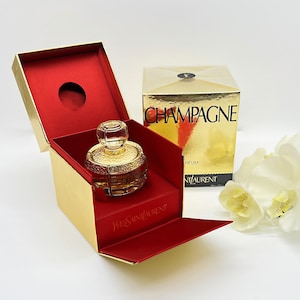 Puede incluir: Una caja dorada y roja con una botella de perfume de vidrio transparente en su interior. La caja está abierta y la botella es visible. La botella tiene una tapa dorada y una etiqueta dorada que dice "Yves Saint Laurent". La caja está etiquetada como "Champagne Parfum".