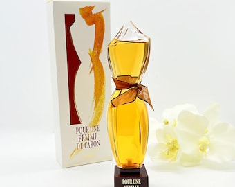 May include: A clear glass bottle of perfume shaped like a woman's dress. The bottle is filled with a golden liquid and has a brown ribbon tied around the neck. The bottle is sitting on a brown base with the words "Pour Une Femme De Caron" written on it. The bottle is in front of a white box with the same text on it.