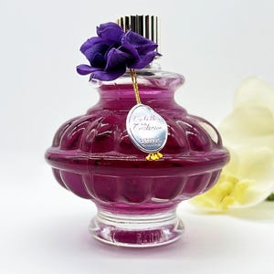 May include: A vintage-style perfume bottle filled with a deep purple liquid. The clear glass bottle has a decorative, ribbed design and is topped with a gold cap and a purple fabric flower. A silver tag hangs from the bottle.