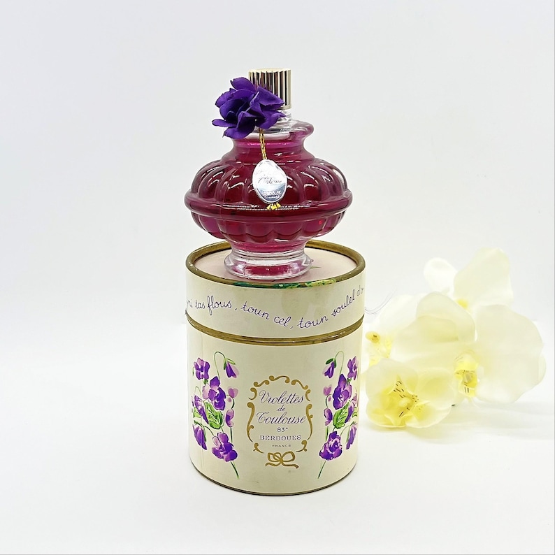 May include: A vintage perfume bottle with a deep red hue, topped with a purple flower and a gold-colored cap. The bottle sits on a cylindrical box adorned with violet illustrations and the text "Violettes de Toulouse". A white flower is in the background.