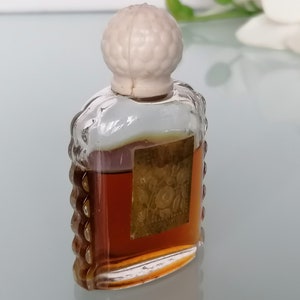 May include: A small, clear glass bottle with a white plastic stopper. The bottle is filled with a brown liquid and has a gold label with a floral design. The label reads "Rose de France".