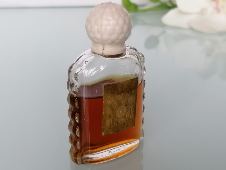 May include: A small, clear glass bottle with a white plastic stopper. The bottle is filled with a brown liquid and has a gold label with a floral design. The label reads "Rose de France".