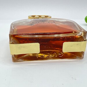 May include: A clear glass bottle of Gucci perfume with a gold cap and a gold label. The bottle is filled with a light brown liquid.