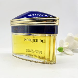 May include: A clear glass bottle of Boucheron Paris Eau de Toilette. The bottle has a gold cap with a blue top and a gold band around the middle. The bottle is filled with a yellow liquid.