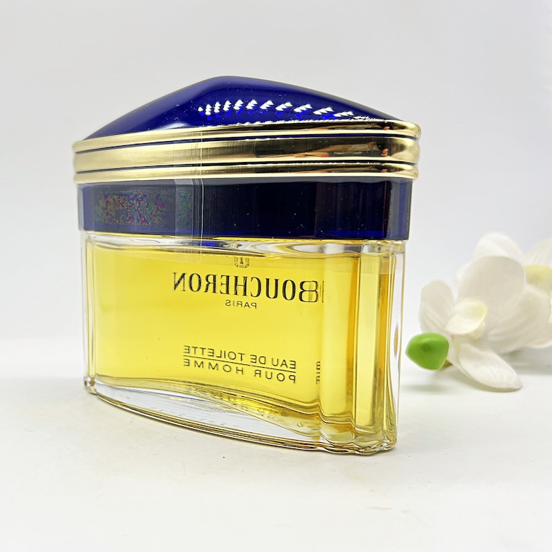 May include: A clear glass bottle of Boucheron Paris Eau de Toilette. The bottle has a gold cap with a blue top and a gold band around the middle. The bottle is filled with a yellow liquid.