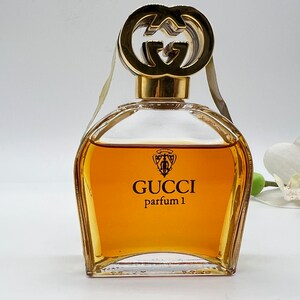 May include: A clear glass bottle of Gucci parfum 1 with a gold double G logo on the stopper. The bottle is filled with a golden liquid.