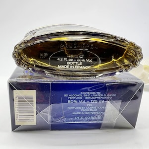 May include: A blue and gold perfume bottle with a label that reads "BOUCHER" and "4.2 Fl. oz - 80% Vol. BOTTLE MADE IN FRANCE". The bottle is sitting on a blue box with white text that includes the ingredients and the text "REF 02-03.06".
