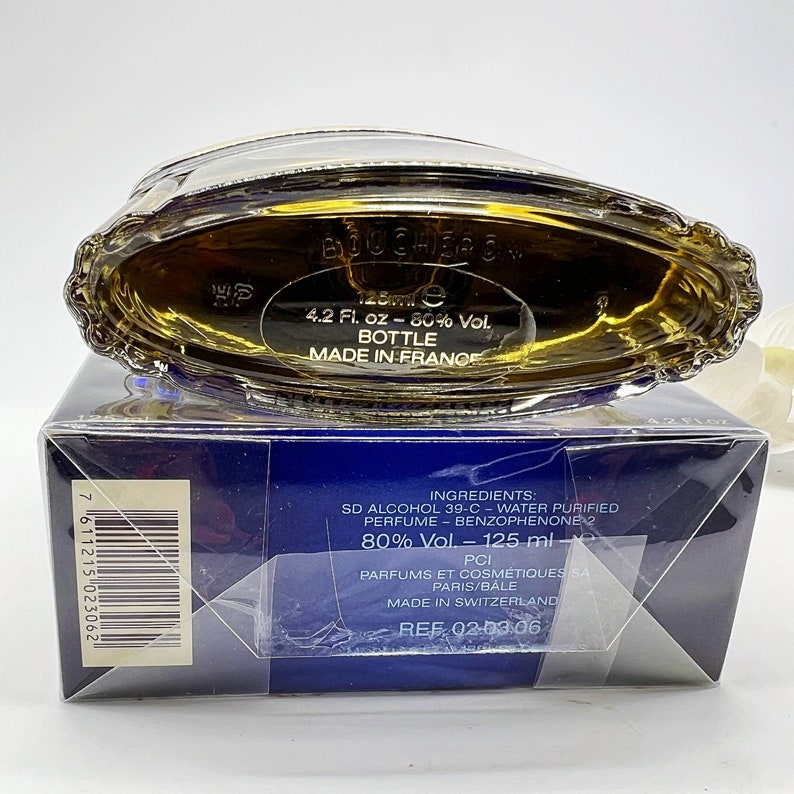 May include: A blue and gold perfume bottle with a label that reads "BOUCHER" and "4.2 Fl. oz - 80% Vol. BOTTLE MADE IN FRANCE". The bottle is sitting on a blue box with white text that includes the ingredients and the text "REF 02-03.06".