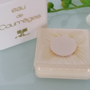 Puede incluir: Una caja cuadrada blanca con el texto "eau de Courr&egrave;ges" y la letra "C" en un c&iacute;rculo. La caja est&aacute; sobre una superficie blanca junto a un peque&ntilde;o recipiente redondo de pl&aacute;stico blanco con una tapa transparente.