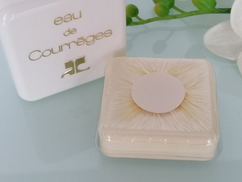 Puede incluir: Una caja cuadrada blanca con el texto "eau de Courr&egrave;ges" y la letra "C" en un c&iacute;rculo. La caja est&aacute; sobre una superficie blanca junto a un peque&ntilde;o recipiente redondo de pl&aacute;stico blanco con una tapa transparente.