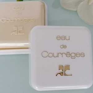 Puede incluir: Dos cajas blancas con letras doradas. La caja superior est&aacute; abierta y muestra una barra de jab&oacute;n blanca con las palabras "eau de Courr&egrave;ges" y la letra "C" en un c&iacute;rculo. La caja inferior est&aacute; cerrada y tiene las palabras "eau de Courr&egrave;ges" y la letra "C" en un c&iacute;rculo.