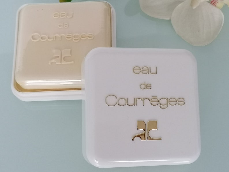 Puede incluir: Dos cajas blancas con letras doradas. La caja superior est&aacute; abierta y muestra una barra de jab&oacute;n blanca con las palabras "eau de Courr&egrave;ges" y la letra "C" en un c&iacute;rculo. La caja inferior est&aacute; cerrada y tiene las palabras "eau de Courr&egrave;ges" y la letra "C" en un c&iacute;rculo.