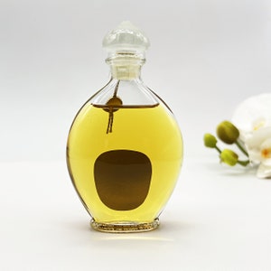 May include: A clear glass bottle with a gold liquid inside. The bottle has a round, bulbous shape and a clear glass stopper with a decorative top. The bottle is sitting on a white surface.