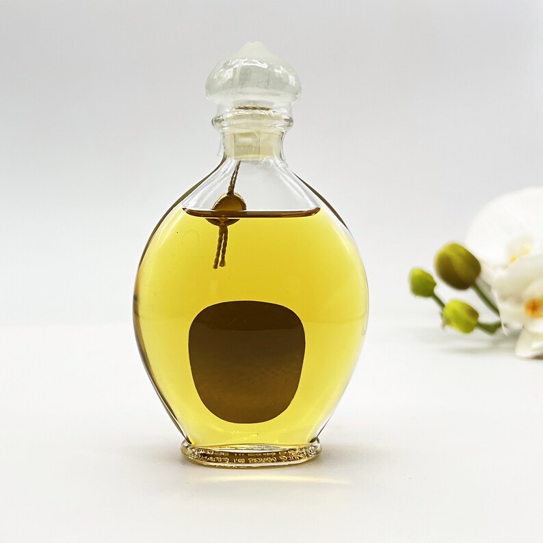 May include: A clear glass bottle with a gold liquid inside. The bottle has a round, bulbous shape and a clear glass stopper with a decorative top. The bottle is sitting on a white surface.