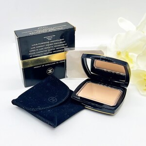 Könnte beinhalten: Ein schwarzes und goldenes Kompaktpuder mit Spiegel, geöffnet, um das Puder zu enthüllen. Eine schwarze Schachtel mit goldenen Akzenten und Text befindet sich hinter dem Kompaktpuder. Ein schwarzes Samtetui und eine Puderquaste sind ebenfalls vorhanden.