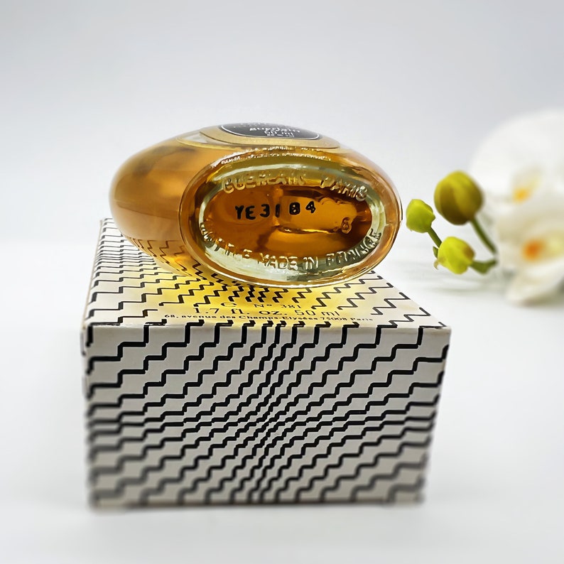 May include: A small, clear glass bottle of perfume with a gold cap. The bottle is sitting on a white box with a black and white geometric pattern. The bottle has a label that says "Guerlain Paris" and "YE 3184".