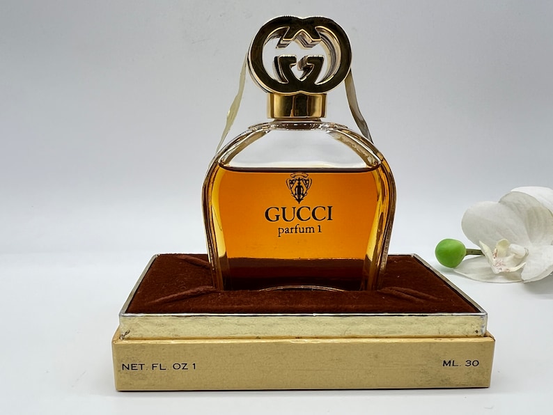 May include: A vintage Gucci perfume bottle in a brown box with gold trim. The bottle is clear glass with a gold cap and the words "GUCCI parfum 1" printed on the front. The box has the words "NET. FL. OZ 1" and "ML 30" printed on the bottom.