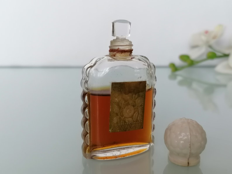 May include: A small, clear glass bottle with a textured design and a brown liquid inside. The bottle has a clear glass stopper and a gold label with a floral design. The bottle is sitting on a white surface next to a white ceramic stopper.