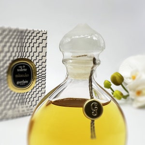 May include: A clear glass bottle of Mitsouko Eau de Toilette by Guerlain. The bottle has a gold cap and a gold tag with the letter "G" on it. The bottle is filled with a yellow liquid.