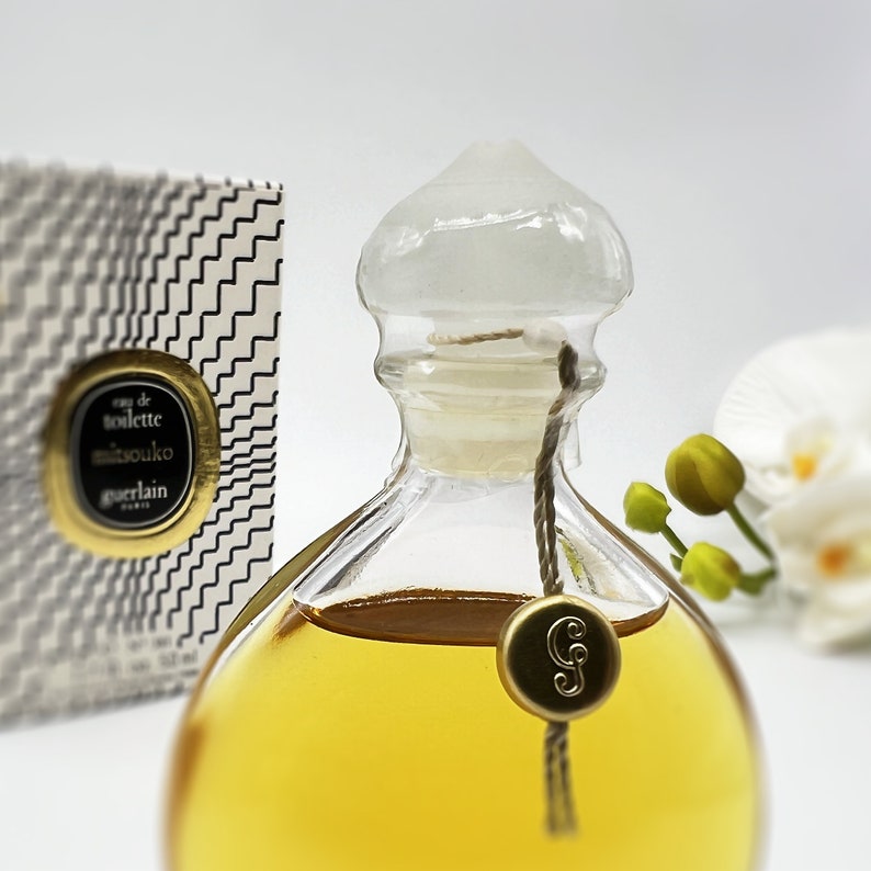 May include: A clear glass bottle of Mitsouko Eau de Toilette by Guerlain. The bottle has a gold cap and a gold tag with the letter "G" on it. The bottle is filled with a yellow liquid.