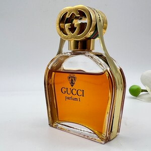 May include: A clear glass bottle of Gucci parfum 1 with a gold double G logo stopper. The bottle is filled with a golden brown liquid.