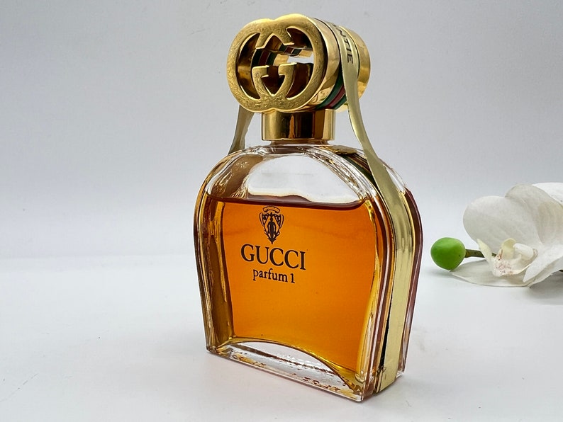 May include: A clear glass bottle of Gucci parfum 1 with a gold double G logo stopper. The bottle is filled with a golden brown liquid.