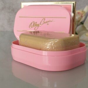 May include: A pink soap dish with a gold trim and the text "Olga Cassini" in gold lettering. The dish contains a wrapped bar of soap.