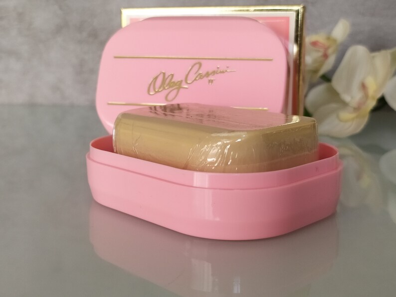 May include: A pink soap dish with a gold trim and the text "Olga Cassini" in gold lettering. The dish contains a wrapped bar of soap.