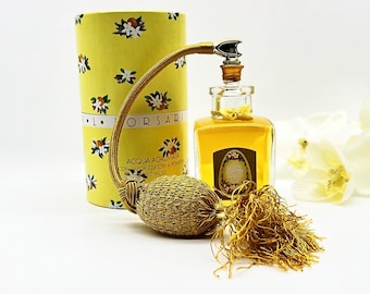 Puede incluir: Un frasco de perfume vintage con un atomizador y borla dorados, junto a una caja cilíndrica amarilla. El frasco está lleno de un líquido dorado y tiene una etiqueta decorativa. La caja y el frasco están decorados con diseños florales blancos.