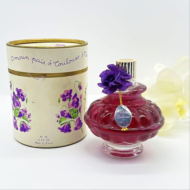 May include: A vintage perfume bottle with a purple flower on top, next to a cylindrical box. The bottle is a deep red color with a silver cap and a small tag. The box has a floral design with the text "Omoun pais, o Coulouse, ol" and "N&deg; 55 4 1/2 OZ Made in France".