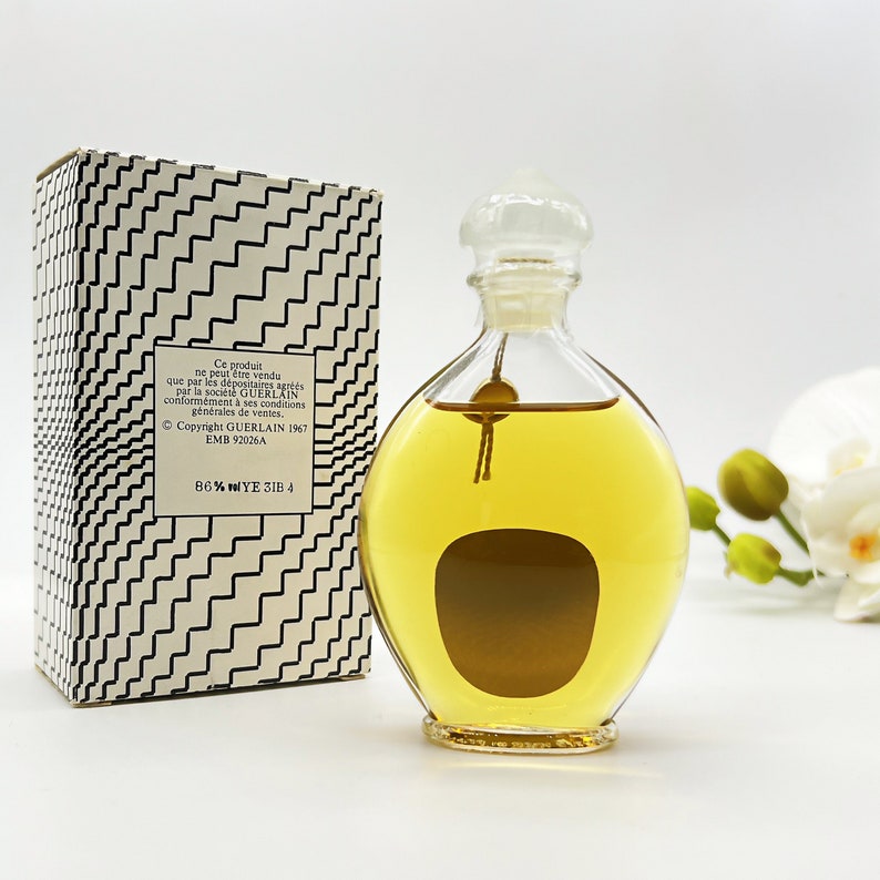 May include: A clear glass bottle of perfume with a gold cap and a brown label. The bottle is sitting on a white surface. The box is white with black geometric patterns and text that reads "Ce produit ne peut &ecirc;tre vendu que par les d&eacute;positaires agr&eacute;&eacute;s par la soci&eacute;t&eacute; GUERLAIN conform&eacute;ment &agrave; ses conditions g&eacute;n&eacute;rales de ventes, Copyright GUERLAIN 1967 EMB 92026A 86% WYE 3IB 4".