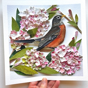 以下が含まれることがあります： ピンクと白の花の枝に止まっているコマドリの紙切り細工。鳥は黒、グレー、オレンジの紙で詳細に作られています。花はピンクと白の紙で、緑の葉が付いています。