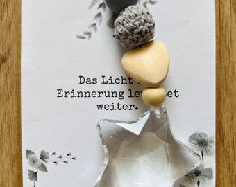 May include: A greeting card with the German text "Das Licht, Erinnerung leuchtet weiter." The card features a clear star-shaped ornament and a beaded strand with wooden and gray beads. Floral illustrations adorn the card's edges.