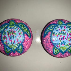 May include: Two pink round knobs with a blue and green sea turtle design. The turtle is outlined in blue and has a pink, green and blue patterned shell.