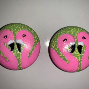 May include: Two round, painted stones feature pink flamingo designs. Each flamingo has a black beak and eye, with a green and yellow patterned border. The background is a light color.