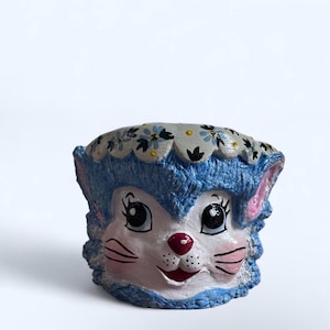 May include: A whimsical, hand-painted planter shaped like a cat's face. The planter features a blue and white color scheme with a floral design around the top. The cat has pink cheeks, a red nose, and a smiling expression.