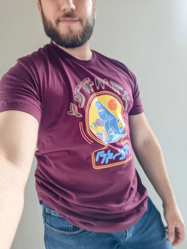 May include: A maroon t-shirt with a graphic of a blue dinosaur in front of a sunset. The dinosaur is standing on a beach with palm trees. The text "F*ck It" is written in Japanese characters below the dinosaur.